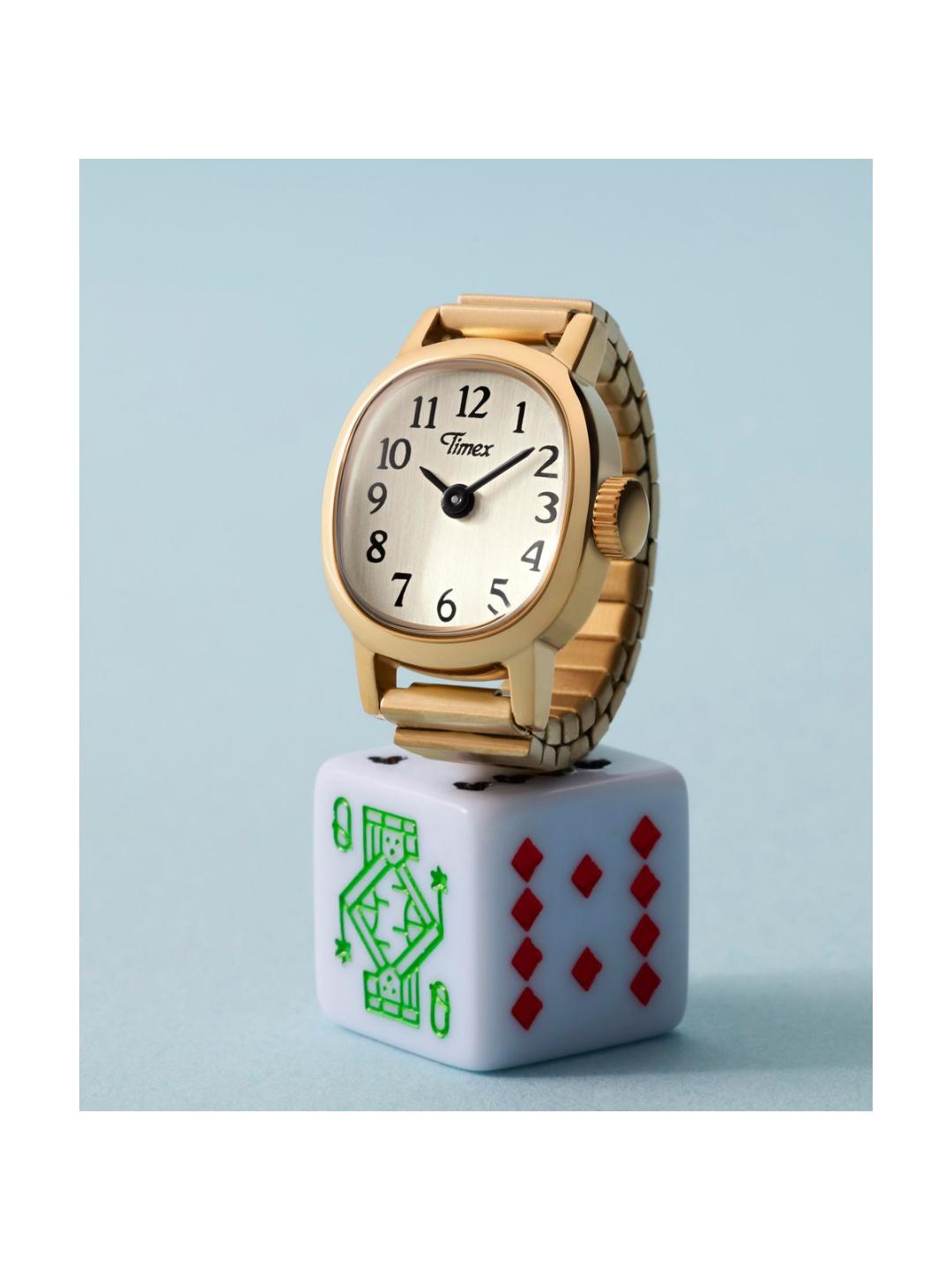 A gold-tone wristwatch with a white dial and black numerals, displayed on top of a white dice block featuring green and red playing card markings, against a pale blue background.