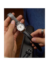 A wristwatch with a silver-toned metal mesh band and a black leather strap, held in a person's hands over a dark case. The watch has a white dial with black Arabic numerals.