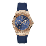 A Guess women's watch featuring a blue dial, rose gold case, and a blue silicone strap. The bezel is embellished with sparkling crystals, and the dial includes Roman numeral XII and sub-dials.